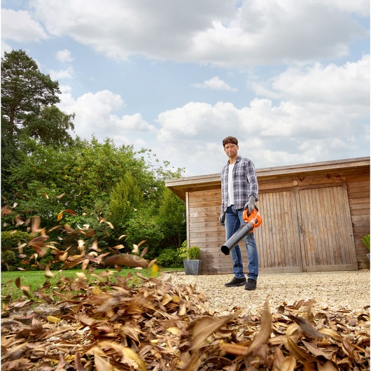 BLACKDECKER BCBL200L leaf blower in use outdoors, blowing leaves on gravel near a wooden shed.