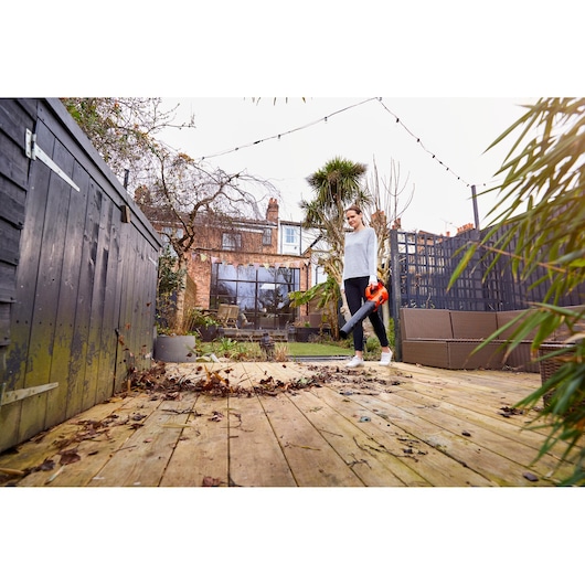 Person using BLACKDECKER BCBL200L cordless leaf blower on wooden deck in backyard garden.