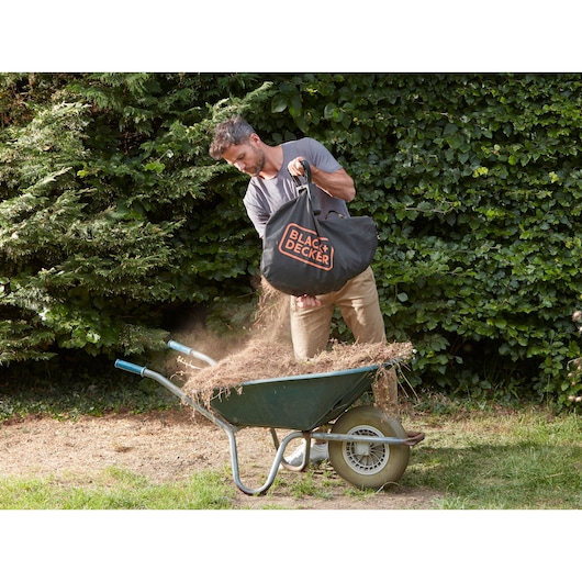 Person emptying BLACK+DECKER BCBLV3620L1 garden vacuum bag into a wheelbarrow outdoors.