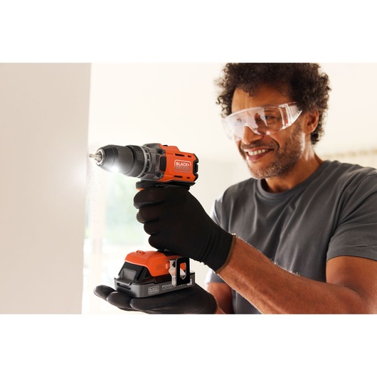 Person using a BLACK+DECKER cordless drill to drill into a wall, wearing gloves and safety glasses.