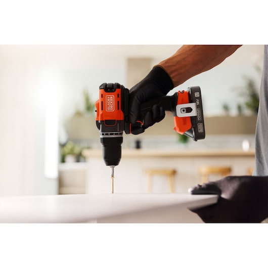 BLACK+DECKER cordless drill in use, drilling into a white surface indoors. User wears black gloves.