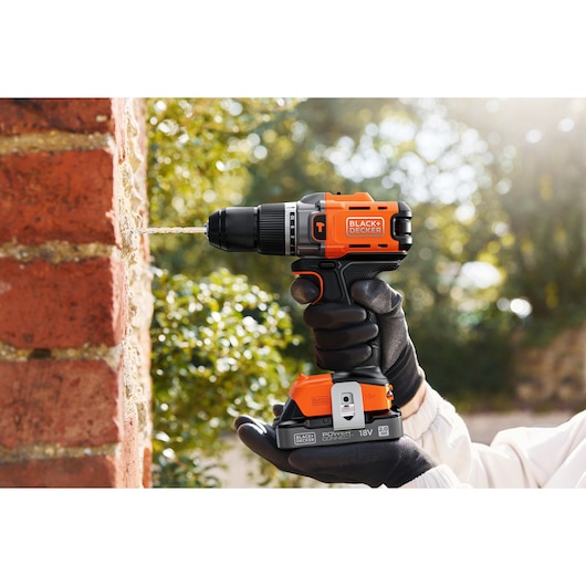 BLACK+DECKER cordless drill used on a brick wall, held by a gloved hand outdoors.