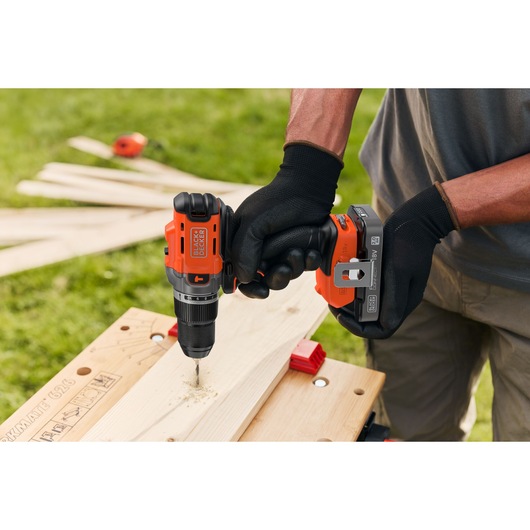 BLACK+DECKER cordless drill in use, drilling into wood on a workbench outdoors.