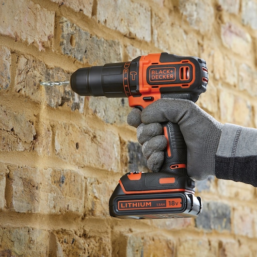 BLACK+DECKER BCD700HTSA cordless drill, black and orange, drilling into brick wall, close-up side view.