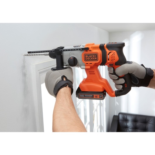 BLACK+DECKER BCD900D1K cordless hammer drill in use, orange and black, drilling into white wall.
