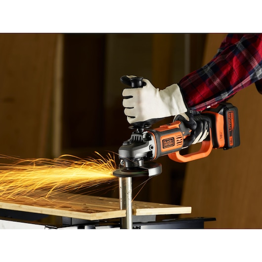 A person wearing work gloves and a plaid shirt is using a cordless BLACK+DECKER angle grinder, creating sparks while grinding a metal pipe.