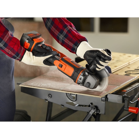 A person wearing gloves and a plaid shirt uses a BLACK+DECKER cordless angle grinder to cut a tile on a workbench. Dust is visible as the tool operates.