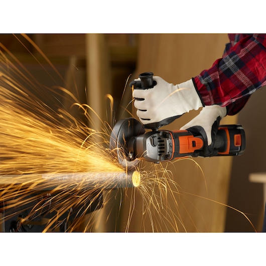 A person wearing gloves and a plaid shirt is using a BLACK+DECKER cordless angle grinder to cut a metal pipe, producing bright sparks.