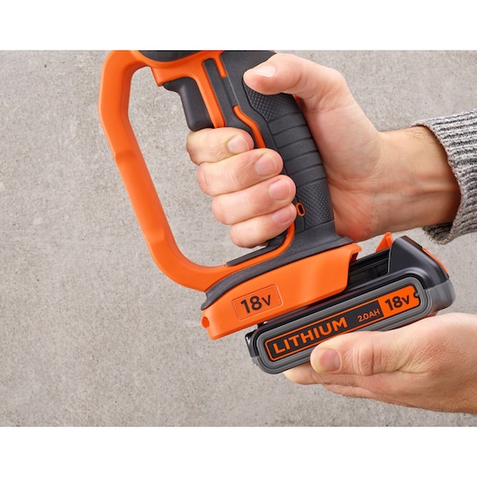 Close-up of a person holding a BLACK+DECKER cordless power tool with an 18V 2.0Ah lithium battery, featuring an orange and black handle against a gray background.