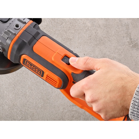 Close-up of a hand pressing the power button on an orange and black BLACK+DECKER cordless power tool against a gray background.