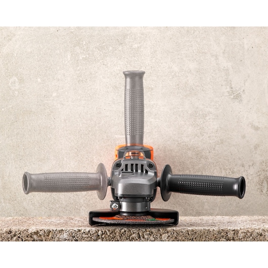 Front view of a BLACK+DECKER angle grinder, positioned on a rough concrete surface against a gray wall, showing three handles and the grinding disc.