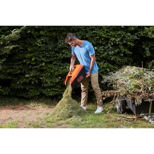BLACKDECKER BCMW33182L2 lawn mower grass box being emptied outdoors near garden waste.