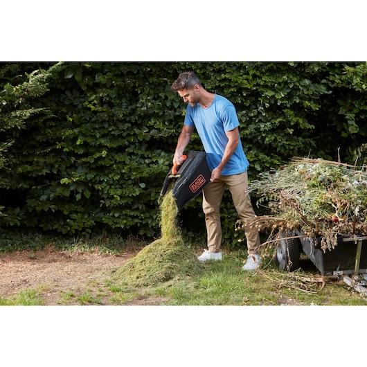 BLACKDECKER BCMW33362L1 lawn mower grass box being emptied onto garden pile outdoors.