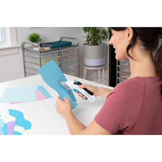 BLACKDECKER BCSC115 cordless cutting tool used to cut blue paper sheets in craft workspace.