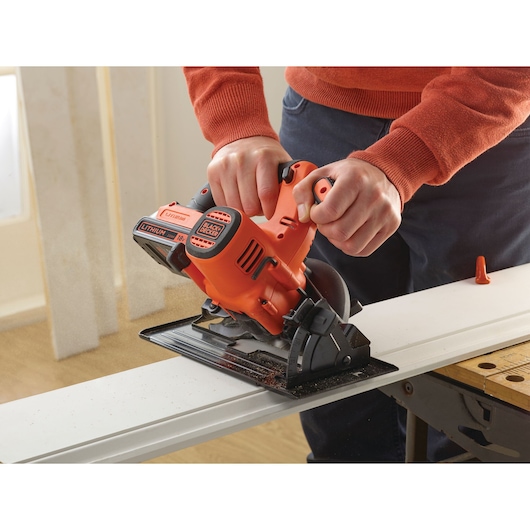 A person using a BLACK+DECKER cordless circular saw to cut a piece of wood, with sawdust visible. The tool is orange and black, and being handled on a workbench indoors.