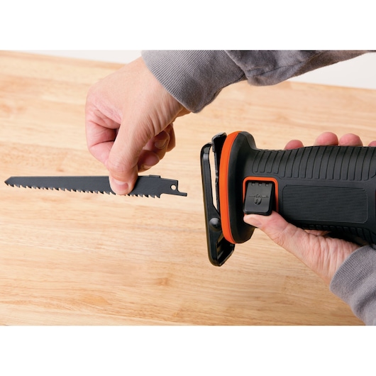A close-up of hands installing a saw blade into a BLACK+DECKER cordless reciprocating saw, positioned over a wooden surface.