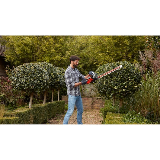 BLACKDECKER BEHTS401 hedge trimmer in use cutting garden shrubs, red and black design, outdoor scene.