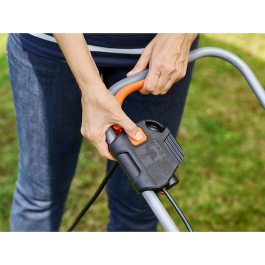 Close-up of BLACK+DECKER BEMW351 lawn mower handle with safety switch and orange button, hands pressing the control.