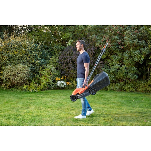 BLACKDECKER BEMW451 lawn mower carried by person in garden with grass and shrubs.