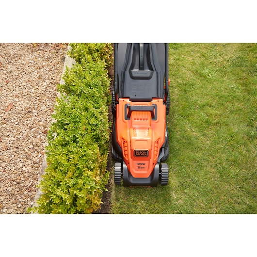 Photo of BLACK+DECKER BEMW471BH electric lawn mower cutting grass, top view, orange and black design beside hedge.
