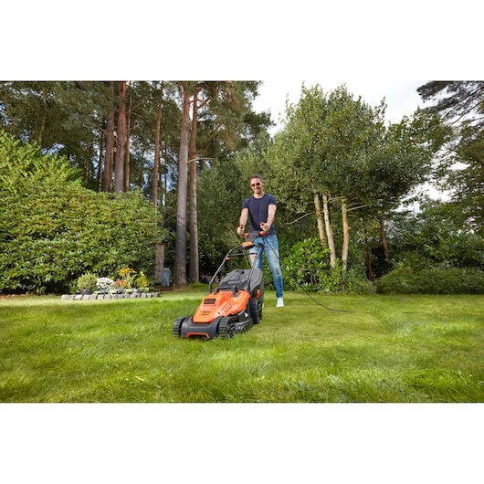 Photo of BLACKDECKER BEMW471ES lawn mower in use cutting grass in a garden with tall trees in the background.