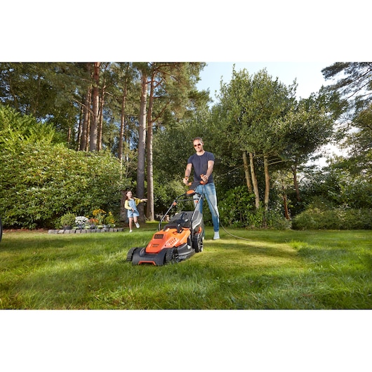 BLACKDECKER BEMW481ES lawn mower in use on grass in a garden with trees and a child in the background.