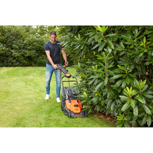 BLACK+DECKER BEMW481ES lawn mower in use on grass near bushes, orange and black design, outdoor setting.