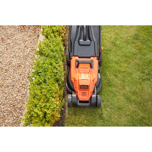 Top view of BLACK+DECKER BEMW481ES lawn mower with orange and black design mowing grass near hedge.