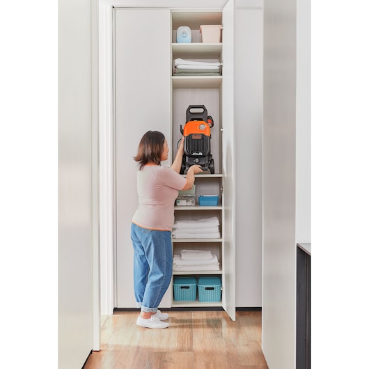 Photo of person storing BLACK+DECKER BEPW2000 pressure washer on a shelf in a white closet with towels and baskets.