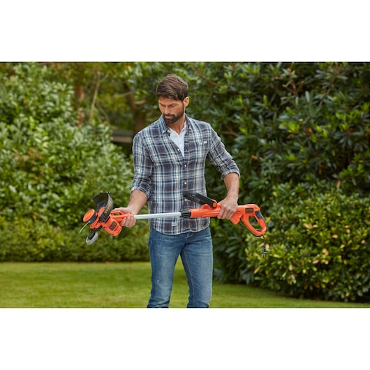 BLACK+DECKER BESTA530 electric grass trimmer with orange and black design held outdoors by a person wearing jeans and plaid shirt.