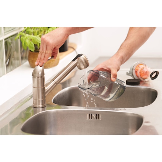 A person cleans a transparent plastic container under a running kitchen faucet, rinsing it over a stainless steel sink. A vacuum cleaner part is placed on the countertop nearby.