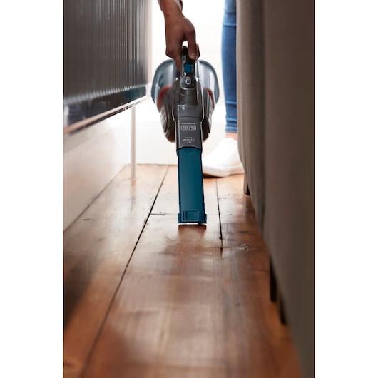 BLACK+DECKER BHHV320B handheld vacuum cleaning narrow space between furniture on wooden floor.