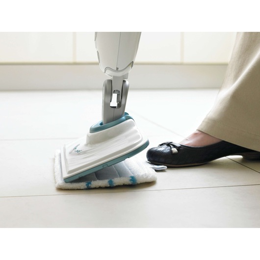 A close-up of a person using a BLACK+DECKER steam mop on a tiled floor. The person's foot is pressing a button on the mop, and the mop head is equipped with a cleaning pad.