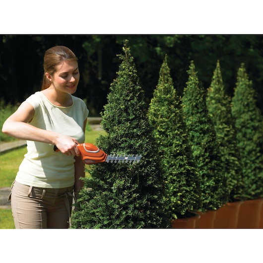 Photo of BLACK+DECKER GSL700 shrub trimmer in use, trimming a potted evergreen bush outdoors.