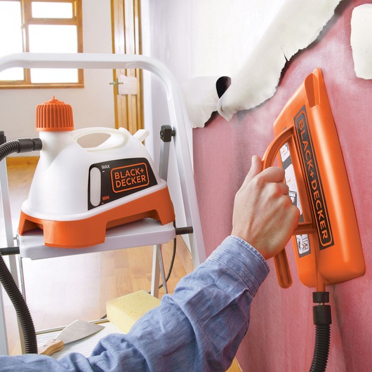 BLACK+DECKER KX3300T wallpaper steamer in use against wall, close-up of orange and white unit and steamer plate.
