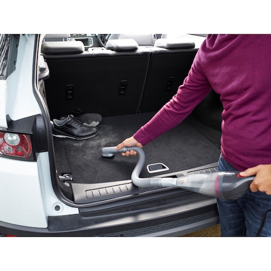 BLACKDECKER NVB12AVA handheld vacuum cleaning car trunk with hose attachment, black shoes in background.