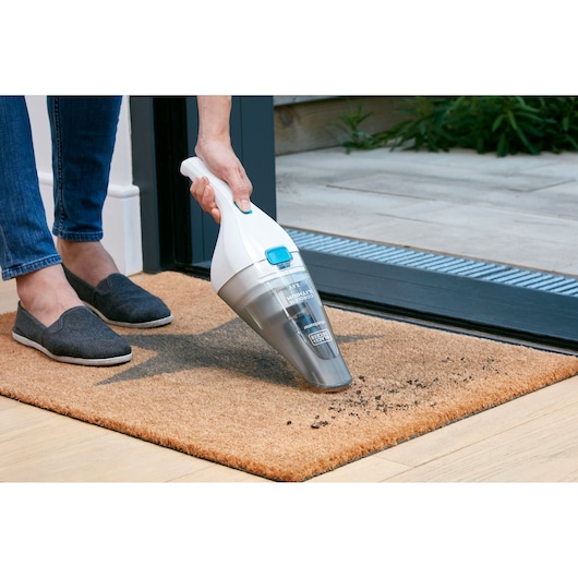 A person using a BLACK+DECKER handheld vacuum cleaner to clean dirt from a doormat placed at an open doorway.