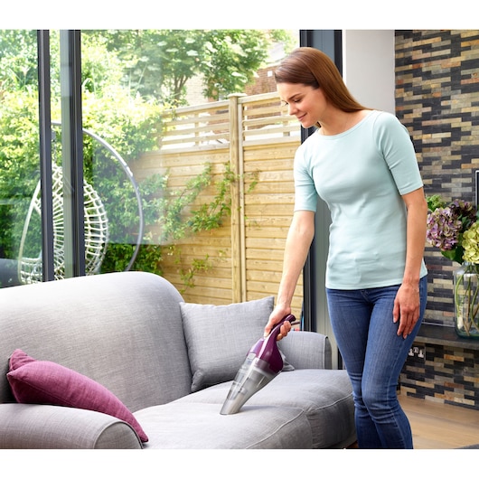 A person using a BLACK+DECKER NVC115W handheld vacuum cleaner to clean a grey fabric sofa in a modern, bright living room with a garden visible outside the window.