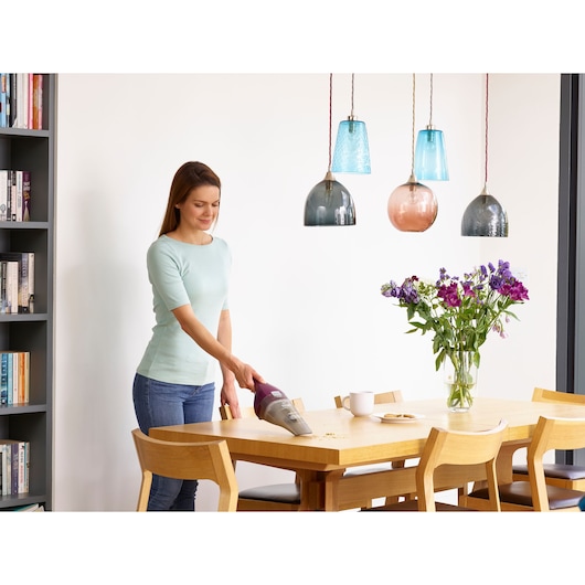 A person using a BLACK+DECKER handheld vacuum cleaner to clean crumbs from a wooden dining table in a modern, well-lit room. There are flowers in a vase and several chairs around the table.