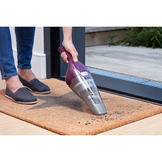 A person using a handheld BLACK+DECKER cordless vacuum cleaner to clean dirt from a doormat near a doorway.