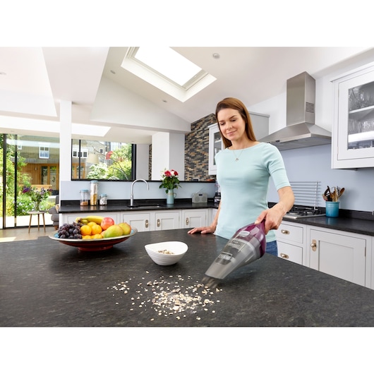 A person using a BLACK+DECKER handheld vacuum cleaner, model NVC115W, to clean spilled cereal on a kitchen countertop. There is a bowl of fruit and a white bowl with cereal nearby. The kitchen is modern and well-lit, with large windows and white cabinets.