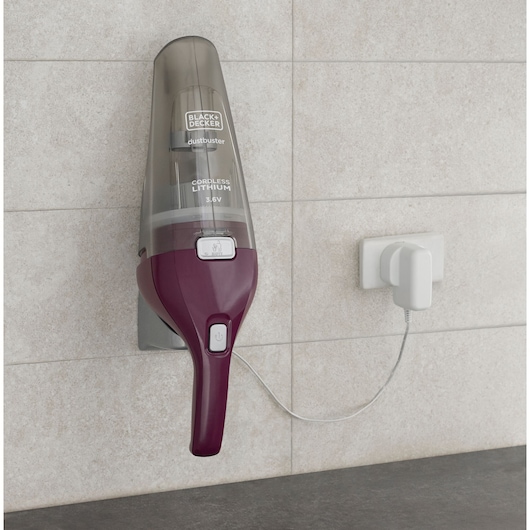 A BLACK+DECKER Dustbuster cordless lithium handheld vacuum, model NVC115W, mounted on a wall and charging via a power outlet, on a tiled background.