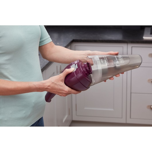 Person holding and emptying a handheld, cordless vacuum cleaner with a purple body in a kitchen setting.