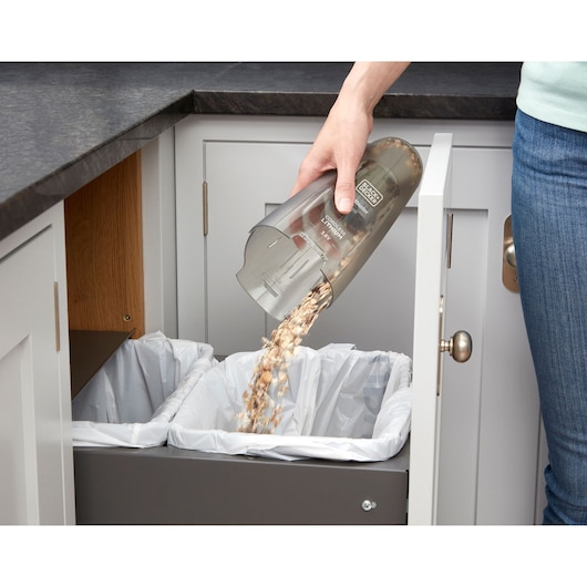A person empties the contents of a BLACK+DECKER handheld vacuum cleaner into a trash bin lined with a plastic bag inside a kitchen cabinet.