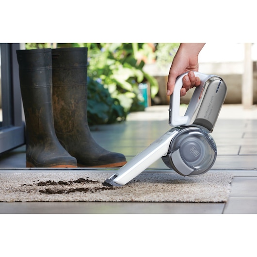 A handheld BLACK+DECKER vacuum cleaner is being used to clean dirt from a rug. A pair of muddy boots is placed near the rug, next to a doorway.
