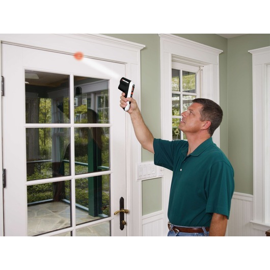 Photo of person using BLACKDECKER TLD100 thermal leak detector near a white glass door indoors.