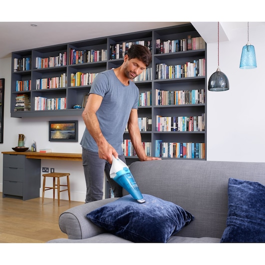 A person using a handheld BLUE BLACK+DECKER vacuum cleaner to clean a cushion on a grey sofa in a modern living room with bookshelves in the background.