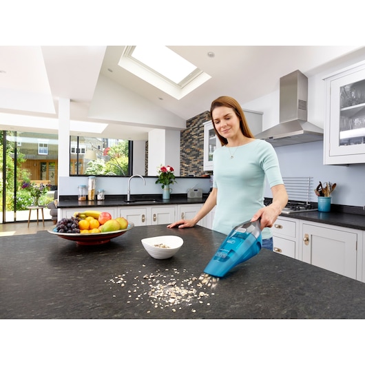 A person using a blue BLACK+DECKER handheld vacuum cleaner (model WDC215WA) to clean spilled oats from a kitchen countertop. The kitchen is modern and bright, with fruit placed in a bowl and a window showing a garden outside.