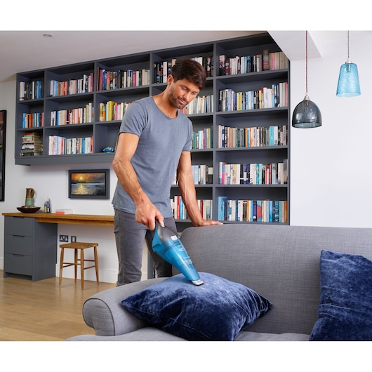 A person using a BLACK+DECKER handheld vacuum cleaner (model WDC215WA) to clean a blue cushion on a grey sofa in a modern living room with bookshelves and pendant lights.