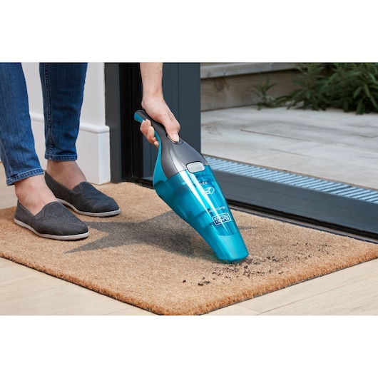 A person uses a blue and grey handheld vacuum cleaner to remove dirt from a doormat near a doorway.